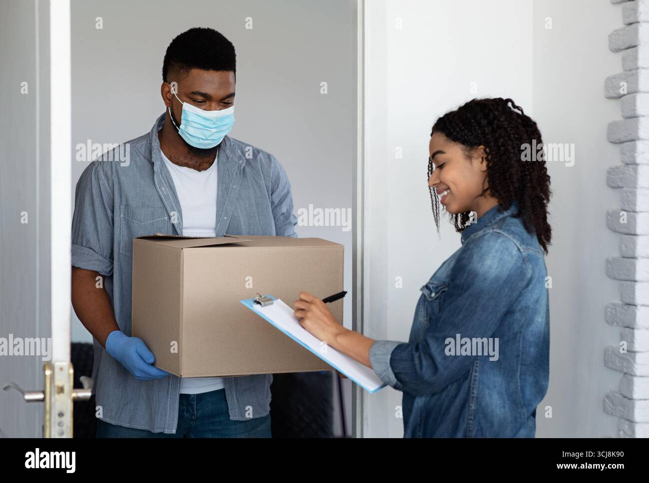 Sichere Lieferung Nach Hause. Schwarze Frau Erhielt Parcel Von Courier In Medizinischer Maske Stockfoto