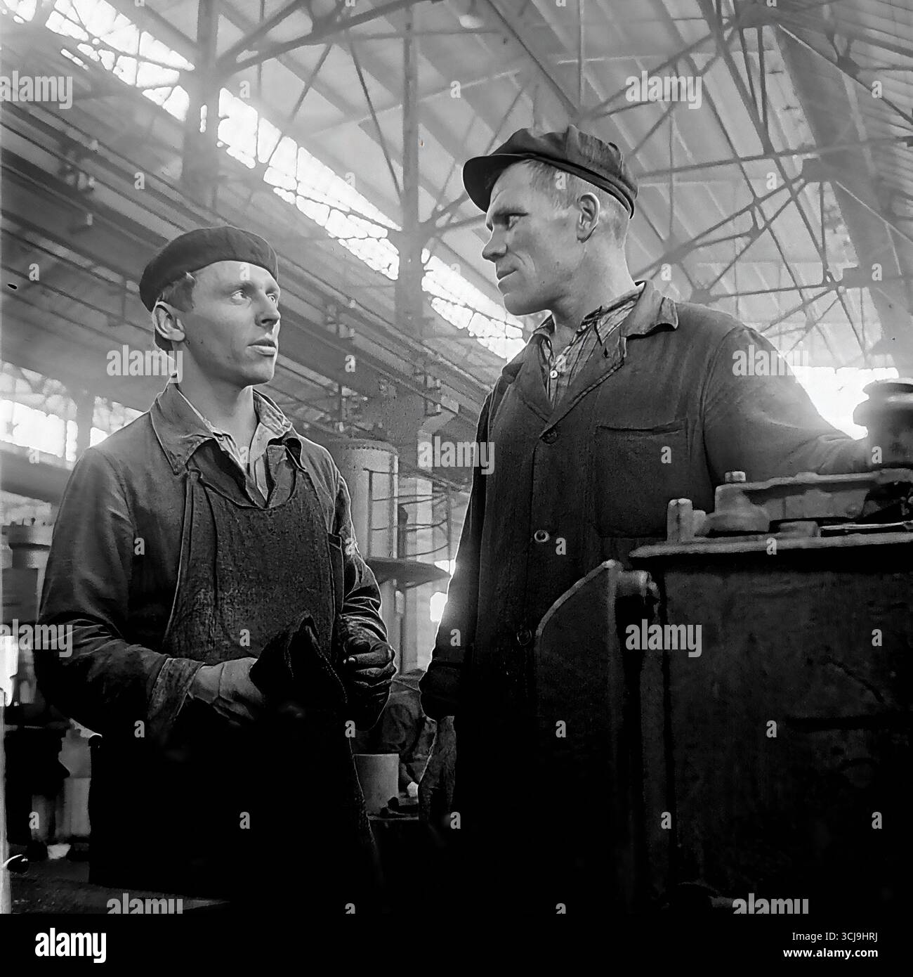 Zwei Arbeitergenossen diskutieren eine Aufgabe an einer Drehmaschine in einer Fabrikwerkstatt in Sloviansk, 1970er Jahre Das Archivfoto vermittelt eine warme Atmosphäre der Freundschaft und Mentorität in einem sowjetischen Werkkollektiv. Diese Szene friedlicher, kreativer Arbeit und menschlicher Beziehungen ist ein Symbol für das Leben im Donbass vor dem Krieg Stockfoto