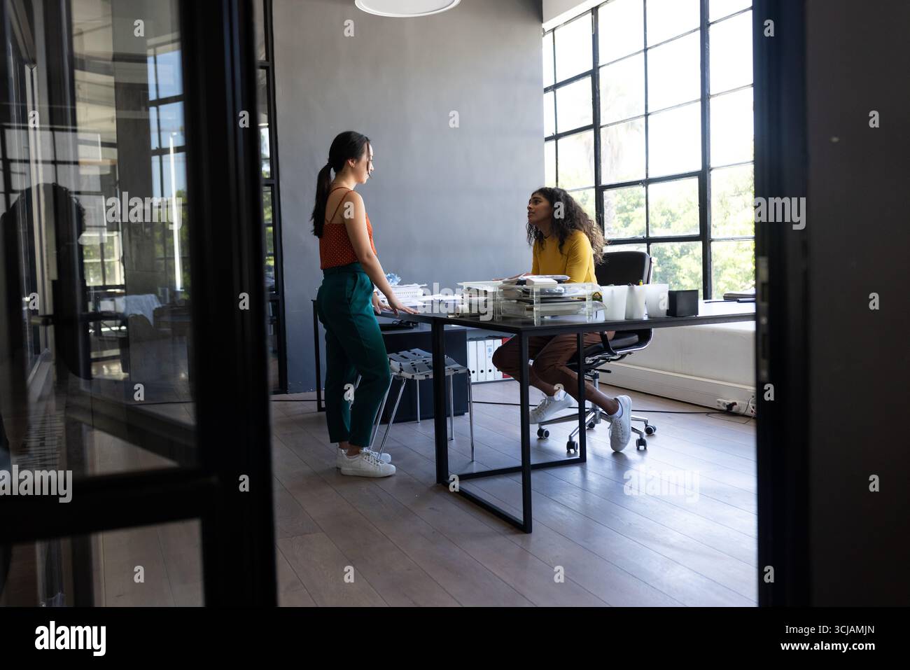Zwei Frauen diskutieren ihre Arbeit am modernen Schreibtisch mit großen Fenstern Stockfoto