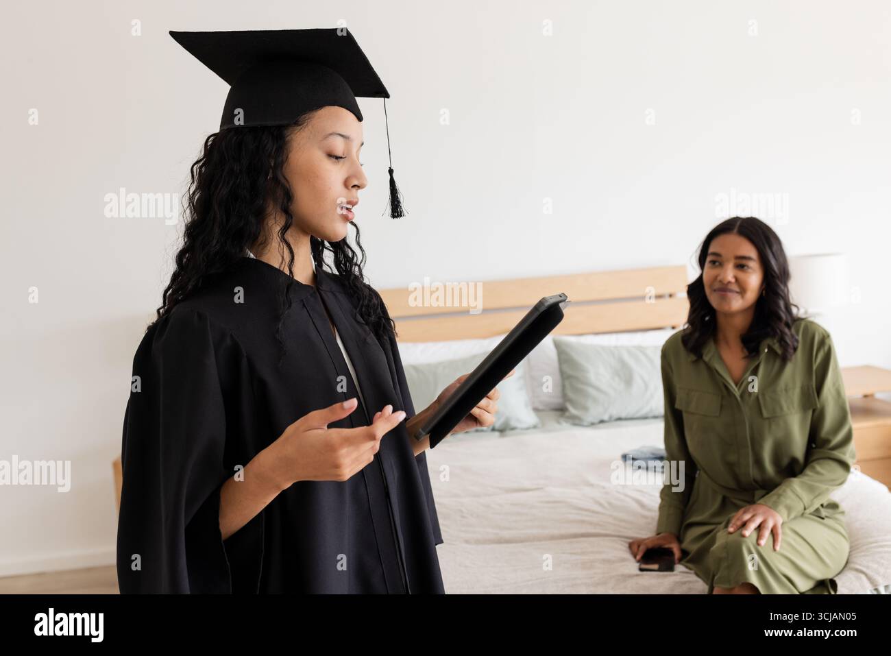 Asiatischer Absolvent in Mütze und Kleid, der Rede zu Hause liest, mit stolzer Mutter, die zusieht Stockfoto
