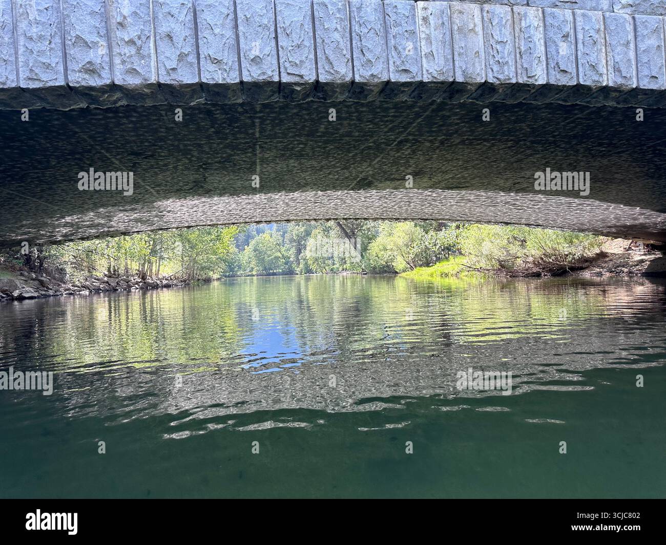 Steinbrücke über einem ruhigen Fluss mit Sonnenlichtreflexionen auf der Unterseite und grünem Wald im Hintergrund. - Smartphone-aufgenommenes Stockfoto