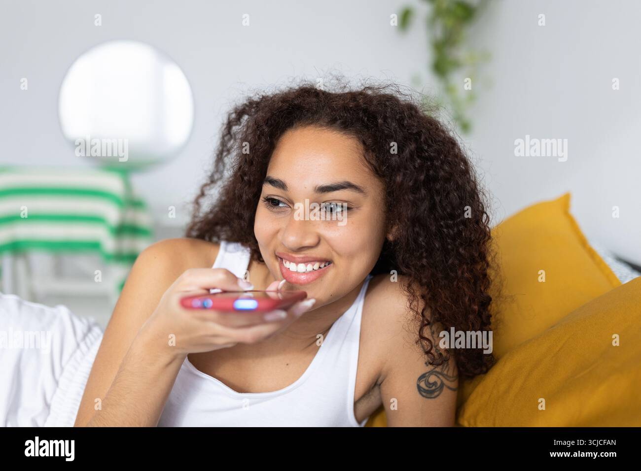 Glückliche lateinamerikanische Frau, die den Sprachassistenten am Smartphone im Bett benutzt Stockfoto