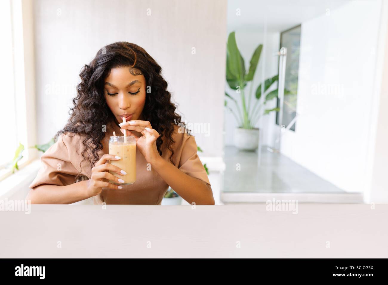 In einem stilvollen, modernen Arbeitsbereich genießt eine Frau einen erfrischenden Eiskaffee. Die Umgebung ist entspannt und zwanglos, mit natürlichem Licht und Grün Stockfoto