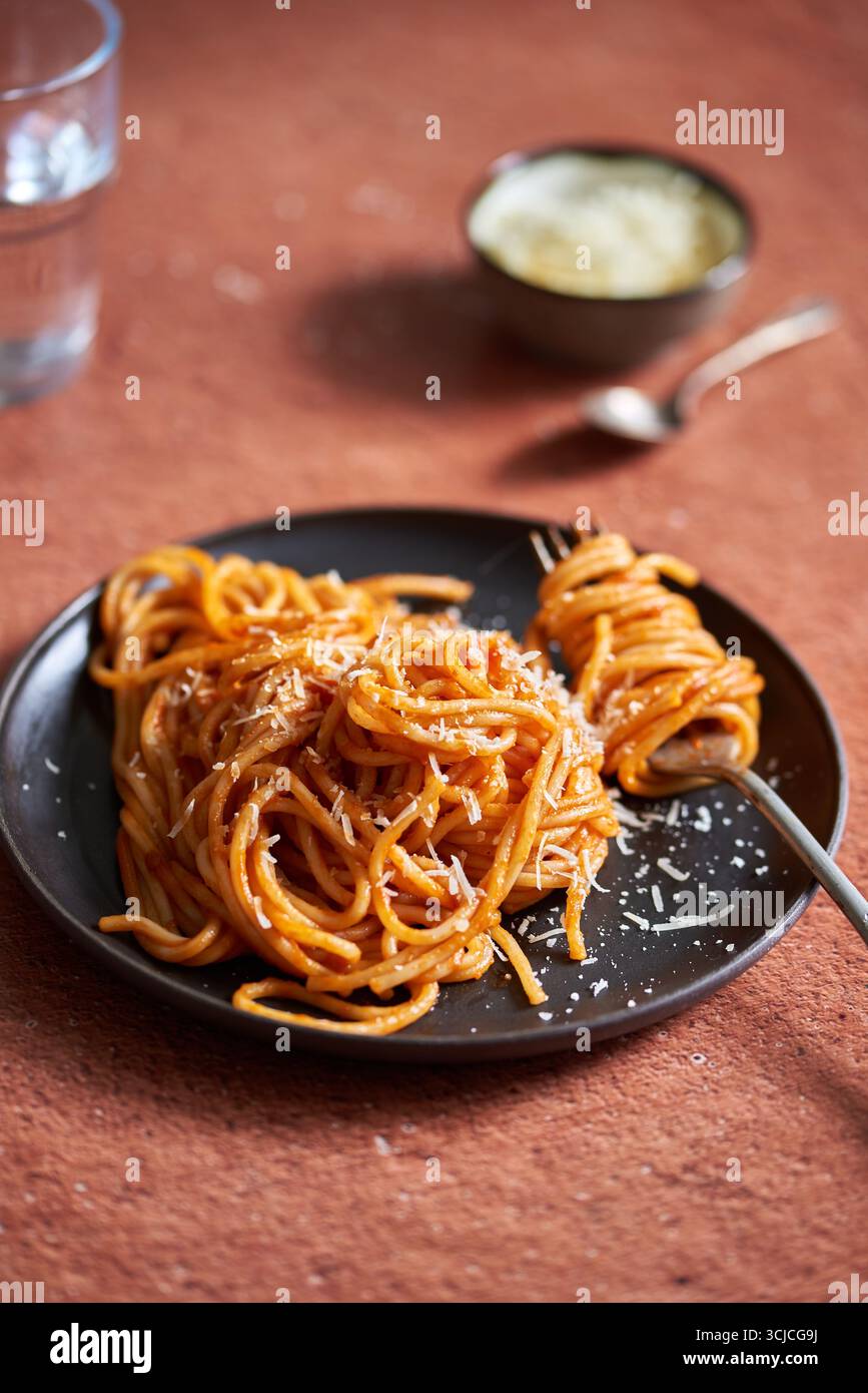 Eine Portion Spaghetti mit reichhaltiger Tomatensauce und geriebenem Parmesankäse, elegant auf einem dunklen Teller präsentiert. Der Hintergrund zeigt ein warmes, t Stockfoto