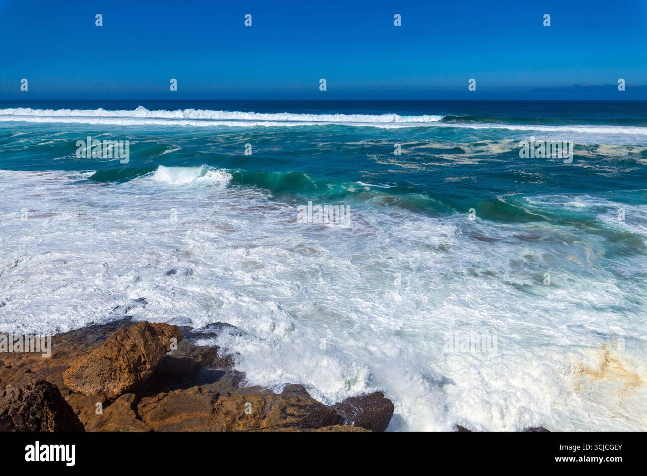 Mächtige Wellen krachen unter einem leuchtend blauen Himmel gegen zerklüftete Küstenfelsen, mit einem entfernten Gebäude an der Klippe mit Blick auf den Ozean Stockfoto