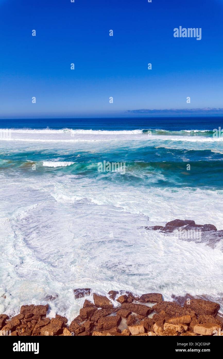 Mächtige Wellen krachen unter einem leuchtend blauen Himmel gegen zerklüftete Küstenfelsen, mit einem entfernten Gebäude an der Klippe mit Blick auf den Ozean Stockfoto