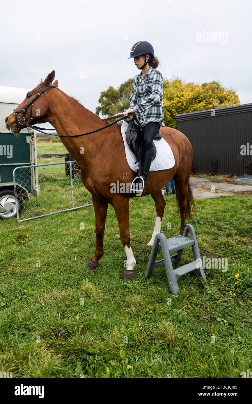 Ein junger Teenager mit Helm reitet auf einem Pferd in der ruhigen Landschaft Neuseelands. Das Bild zeigt die Reiterkultur und den ländlichen Lebensstil, Stockfoto