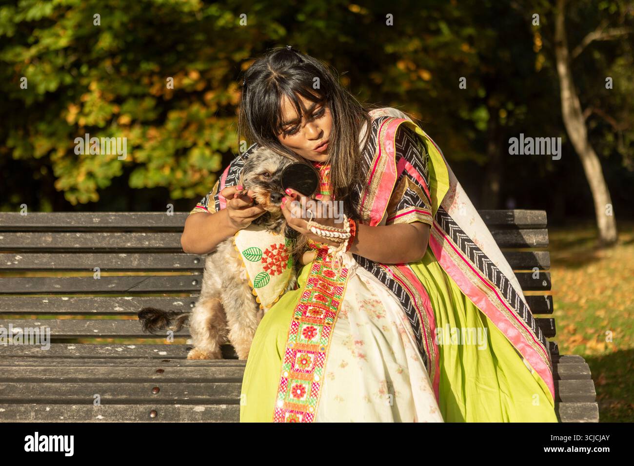 Indische Frau in traditioneller Kleidung, die auf einer Parkbank sitzt und ihren kleinen Hund umschließt, während sie einen sonnigen Herbsttag genießt. Die lebendigen Blätter führen zu einem Krieg Stockfoto