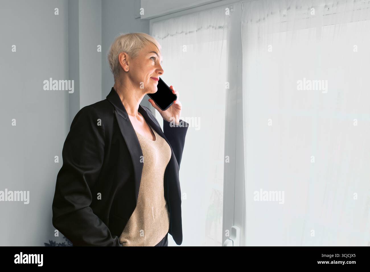 Eine Frau in einem schwarzen Blazer steht an einem Fenster, während sie von ihrem Heimbüro aus arbeitet und ein Smartphone an ihr Ohr hält. Sie strahlt Selbstvertrauen aus und Stockfoto