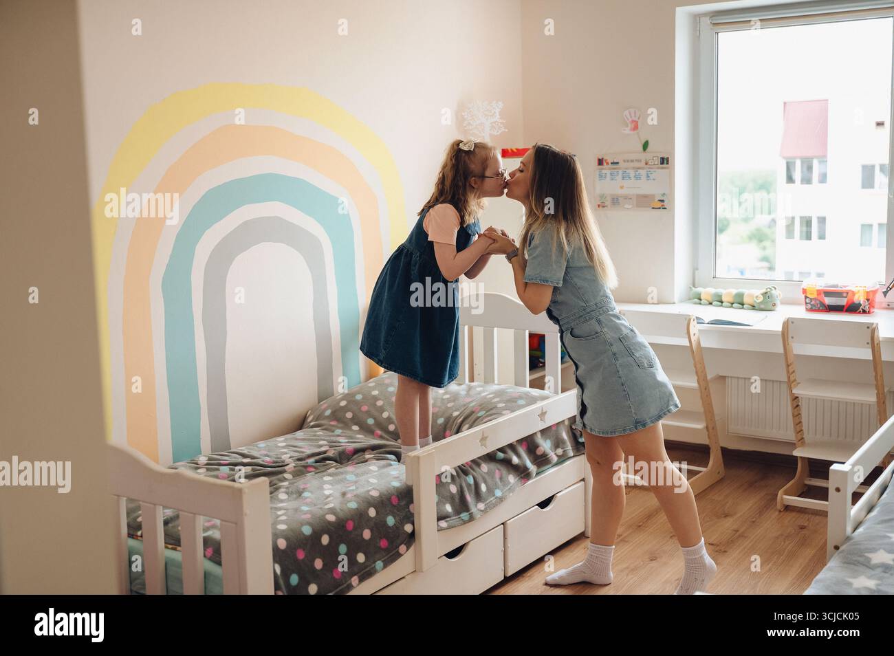 Ein berührender Moment zwischen einer Mutter und ihrer Tochter mit Down-Syndrom, während sie einen liebevollen Kuss teilen, Seite an Seite auf dem Bett stehen in einem gemütlichen, warmen Ambiente Stockfoto
