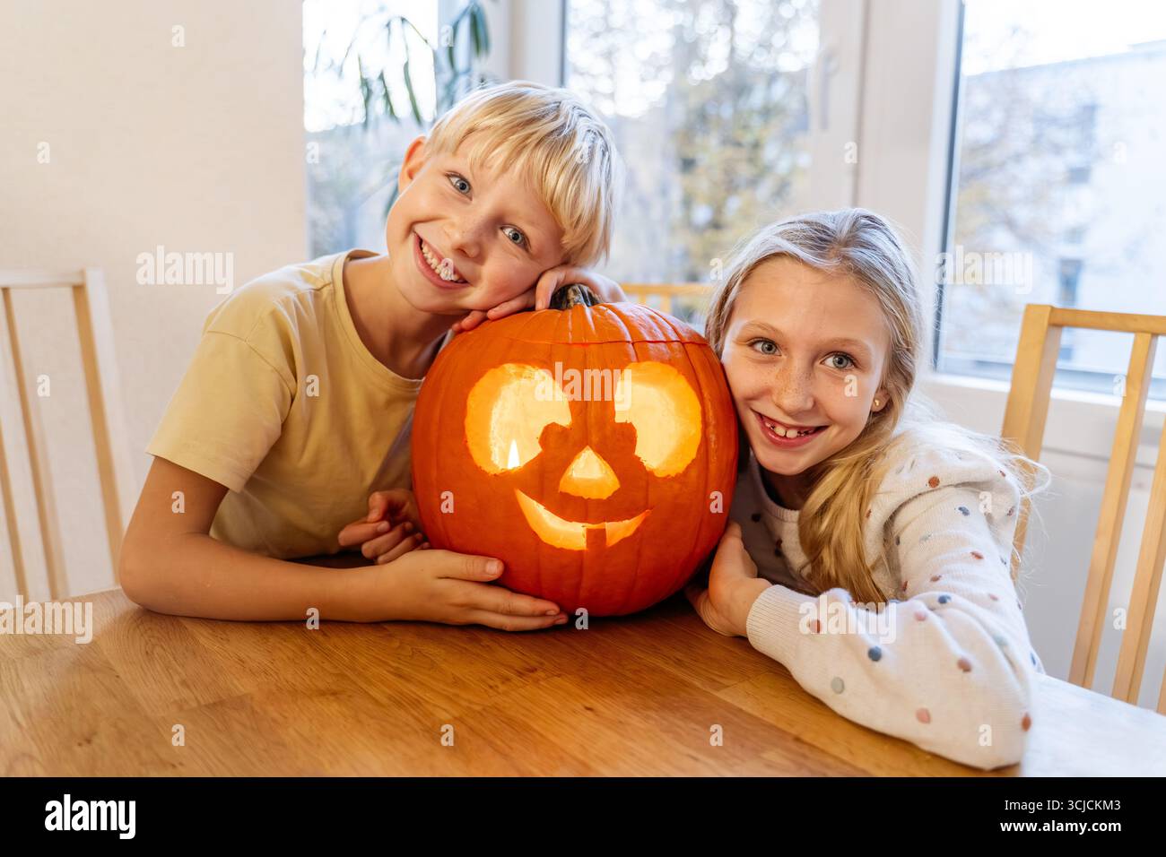 Zwei lächelnde Kinder umarmen zu Hause einen geschnitzten Halloween-Kürbis. Der leuchtende Kürbis sitzt auf einem Tisch und schafft eine warme und festliche Atmosphäre voller Freude und Freude Stockfoto