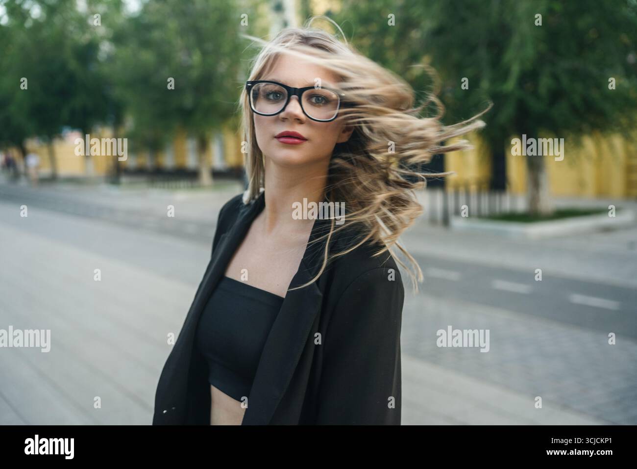 Die junge blonde Frau mit großer schwarzer Brille genießt einen luftigen Tag im Freien, ihr Haar fließt im Wind vor einem städtischen Hintergrund. Sie strahlt Vertrauen aus Stockfoto