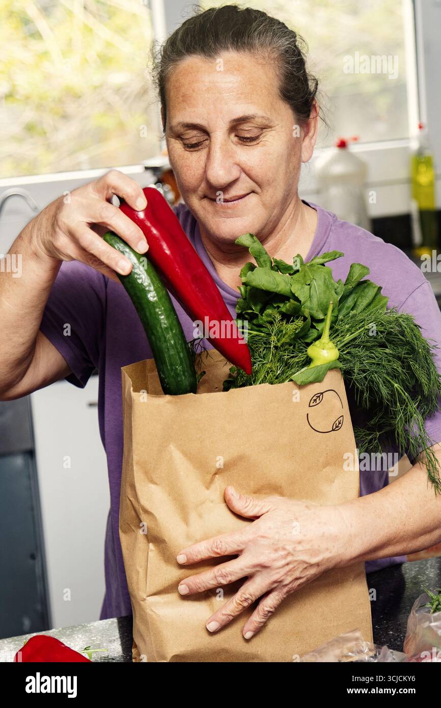 Frau mittleren Alters, die frisches Gemüse in einer Papiertüte in einer gemütlichen Küche anlegt. Sie scheint zufrieden zu sein und betont die Wichtigkeit einer gesunden Ei Stockfoto