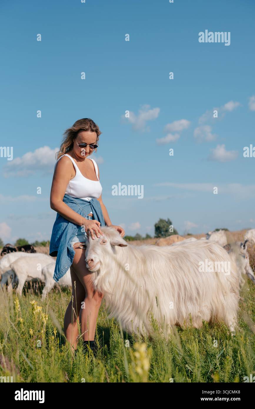 Eine lächelnde Frau mit Sonnenbrille und lässigem Outfit genießt einen sonnigen Tag auf einem Bauernhof, umgeben von Ziegen. Der klare blaue Himmel Stockfoto
