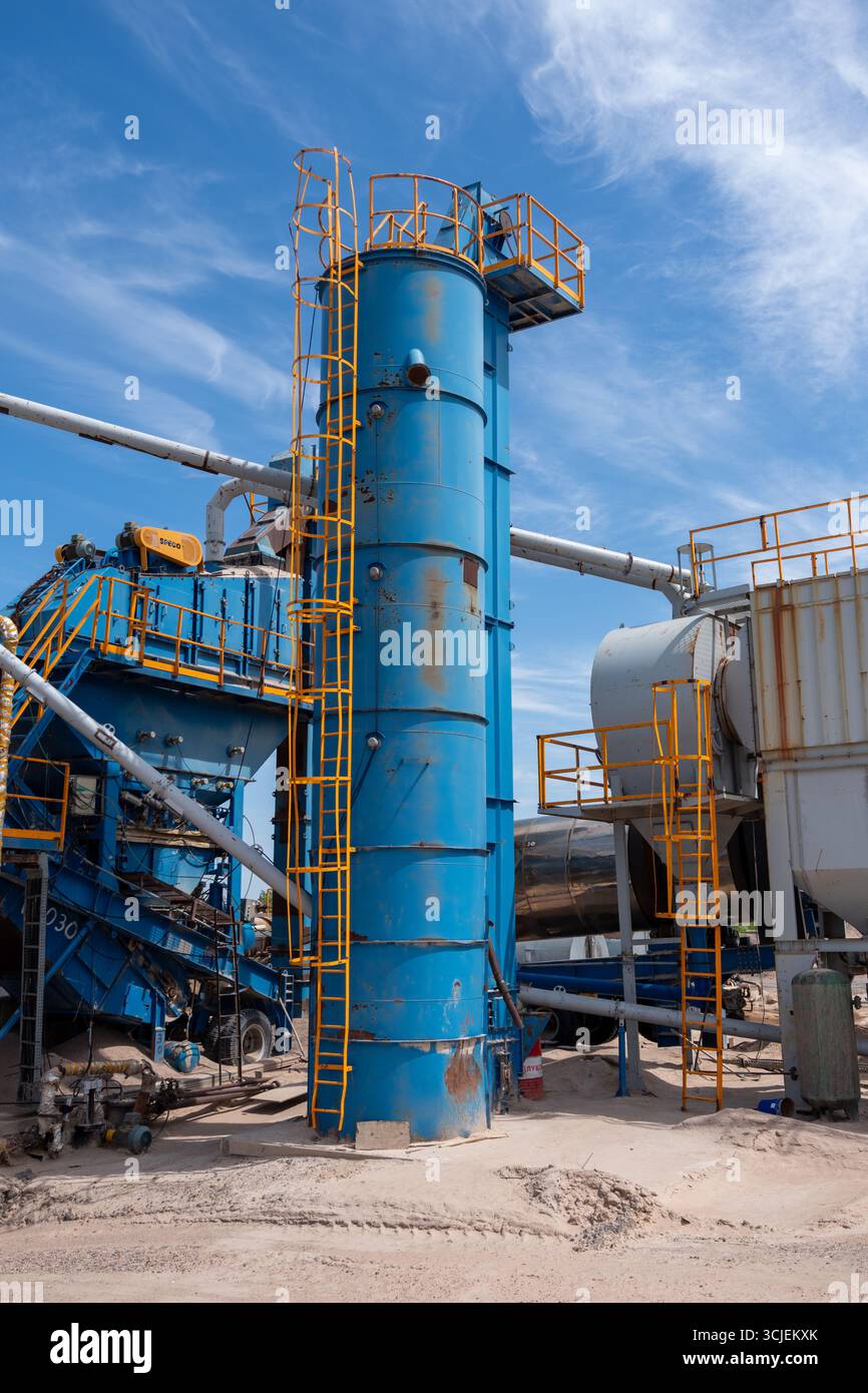 Großes blaues Industriesilo mit gelben Leitern in einer Asphaltmischanlage mit Metallrohren und Maschinen an einem sonnigen Tag Stockfoto