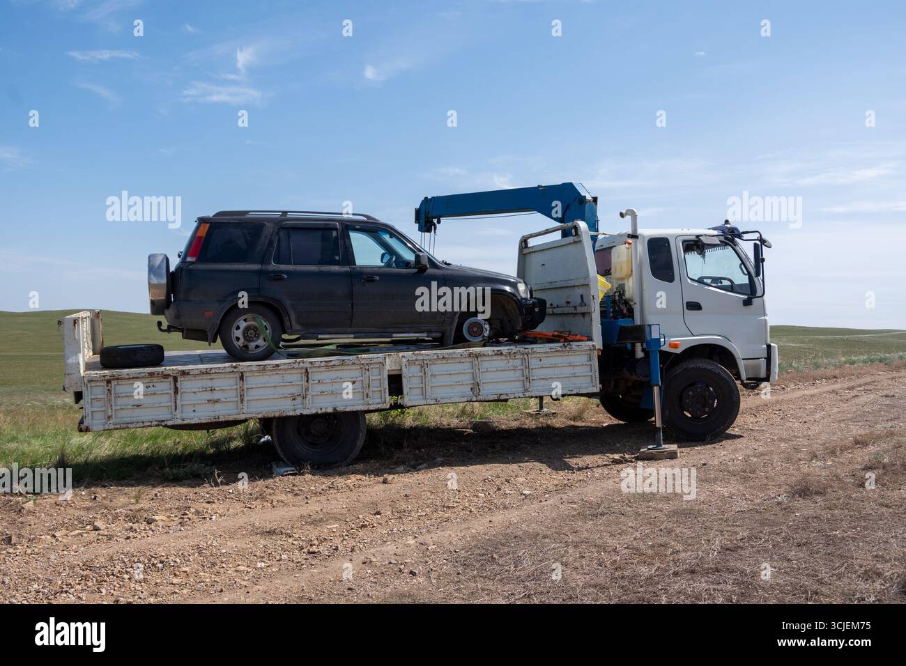 Beschädigtes SUV-Fahrzeug wird mit einem Kran auf einen Tieflader auf unbefestigter Straße, natürlichem Tageslicht, ländlichem Hintergrund, Pannenhilfe-Konzept angehoben Stockfoto