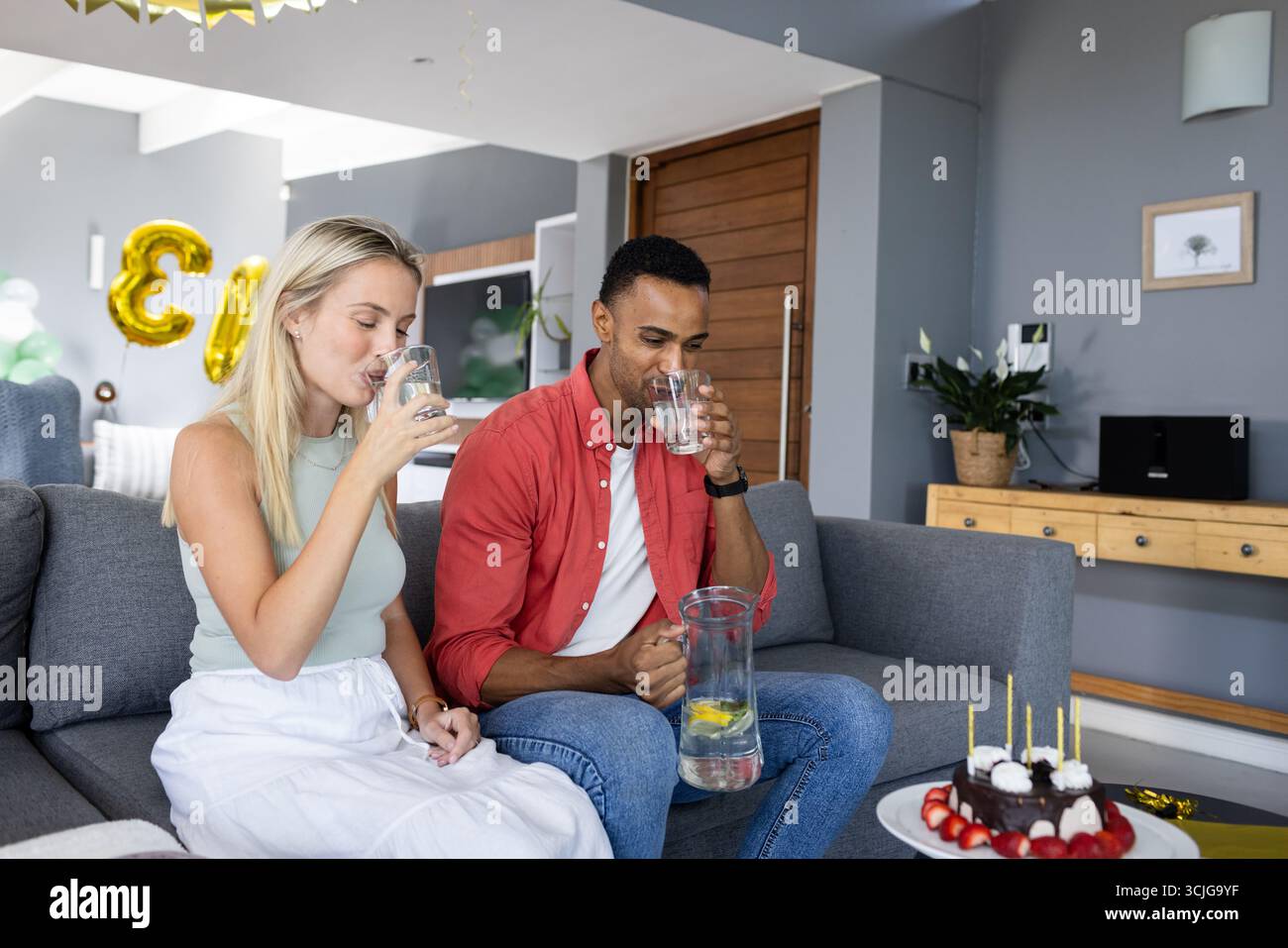 Paare genießen erfrischende Getränke auf dem Sofa mit Kuchen und Ballons in der Nähe Stockfoto