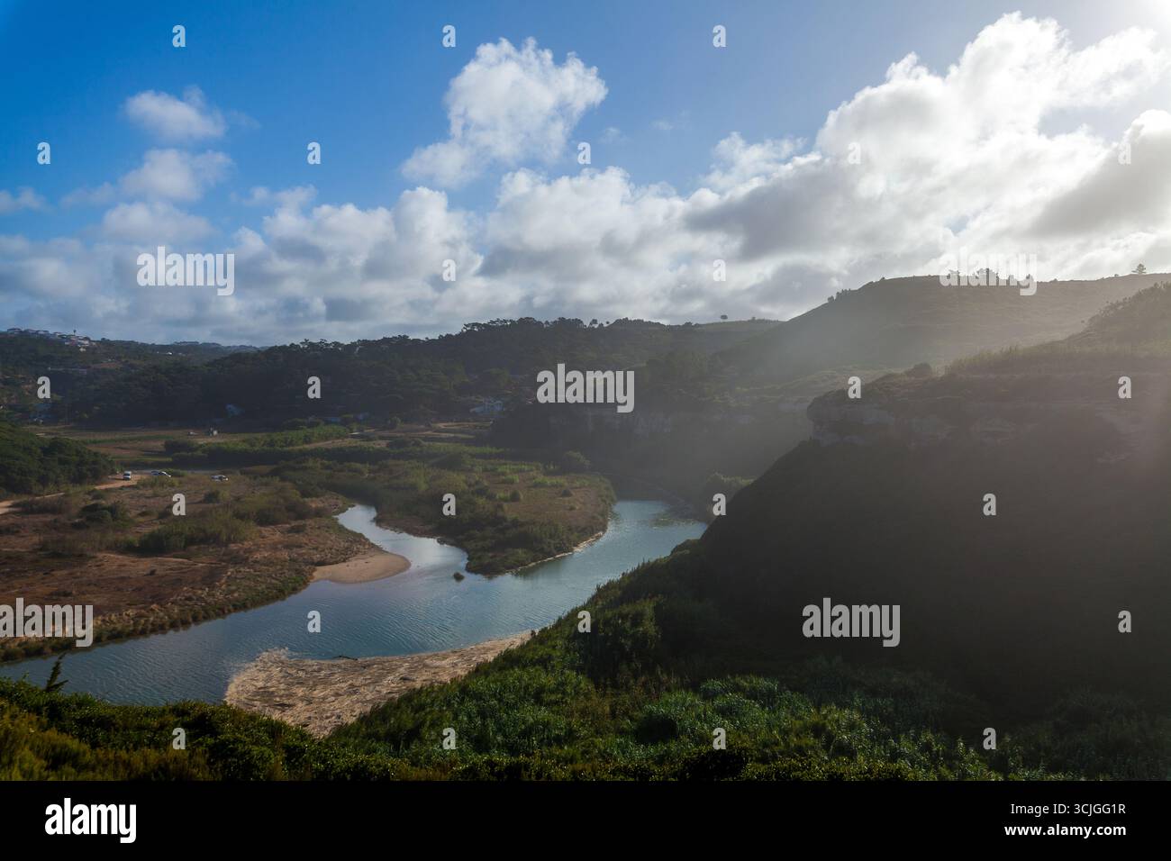 Ein sich windender Fluss fließt durch ein üppiges Tal, umgeben von sanften Hügeln, dichter Vegetation und dramatischen Klippen unter einem hellblauen Himmel mit verstreuten Flächen Stockfoto