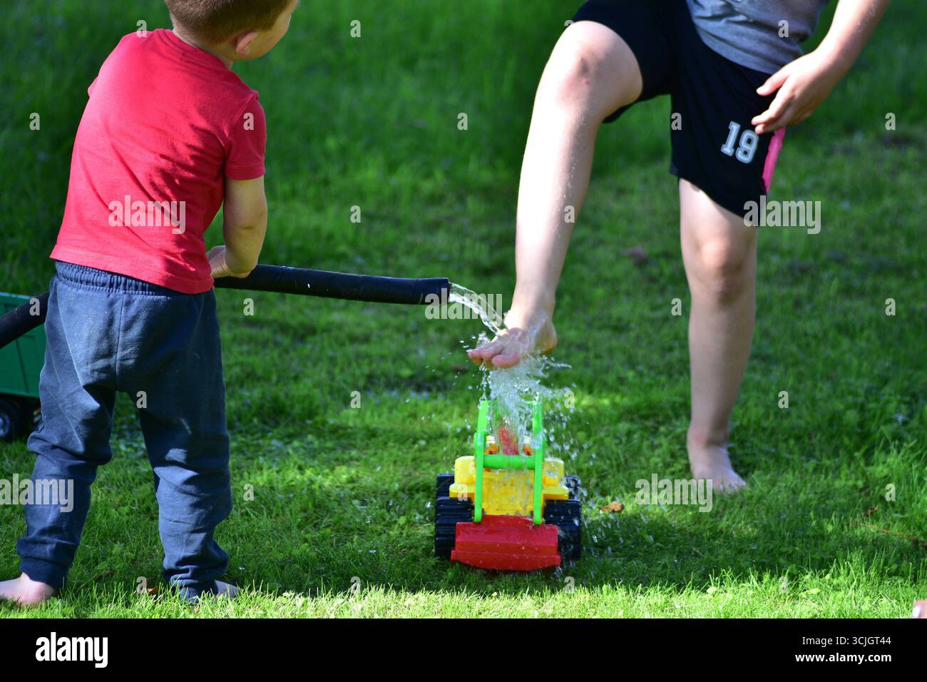 Kinder spielen mit Gartenschlauch und Spielzeugtraktor und spritzen im Sommer Wasser auf Gras. Unterhaltsame Aktivitäten im Freien und Familienurlaub. Stockfoto