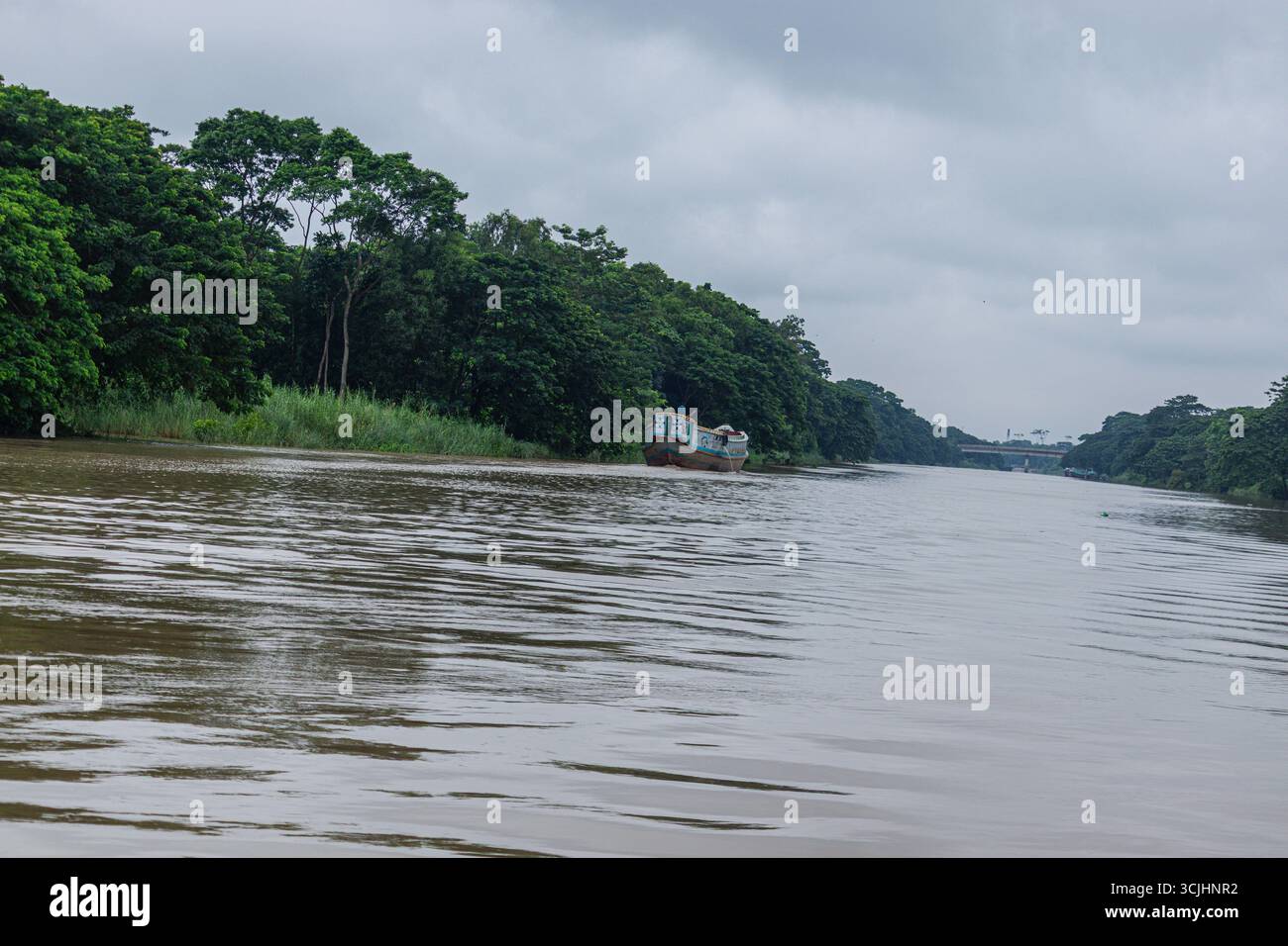 Verblichenes, lackiertes Frachtboot gleitet an grünen Flussinseln unter feuchten, grauen Monsunwolken vorbei Stockfoto
