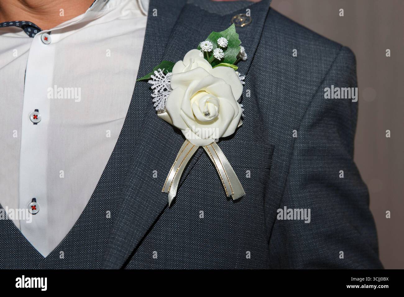 Ein Mann in einem formellen Anzug mit einer weißen, schönen Blumenkuppe. Der Hintergrund ist leicht verschwommen, was auf eine festliche oder feierliche Atmosphäre hindeutet Stockfoto