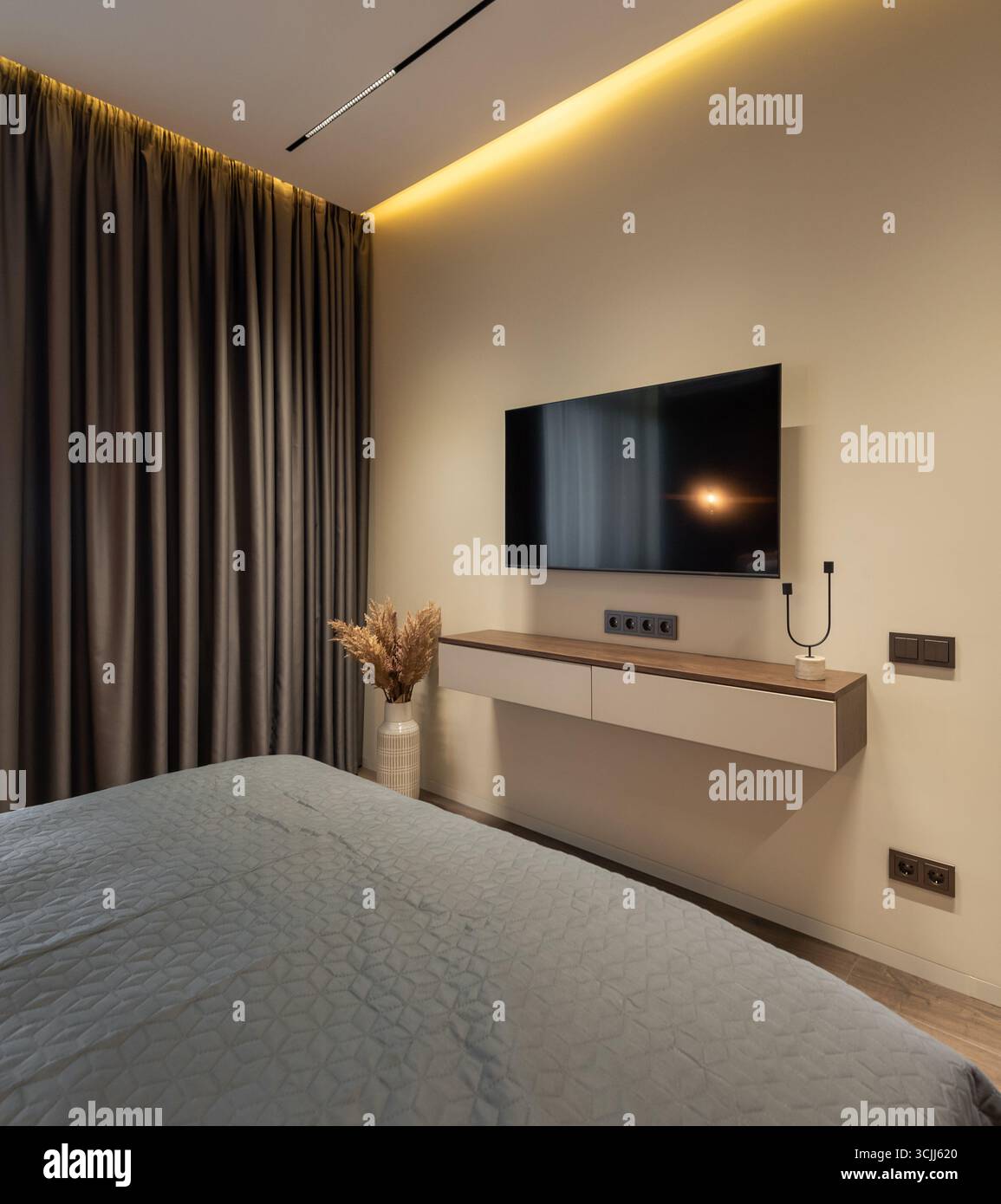 Wandmontierter Fernseher im Schlafzimmer. Großer Flachbildschirm an einer cremebeigen Wand, Zimmer mit beleuchteter Decke und braunem Vorhang. Stockfoto
