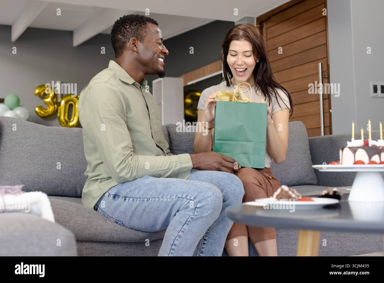 Ein Paar tauscht Geschenke auf dem Sofa aus, feiert Geburtstag mit Kuchen und Ballons Stockfoto