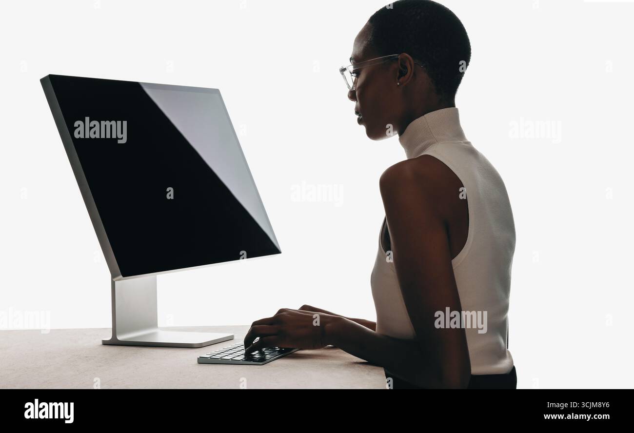 Eine fokussierte Person mit Brille arbeitet an einem schlanken Computer-Setup in einer sauberen, hellen Umgebung, die Professionalität und Modernität verkörpert. Stockfoto