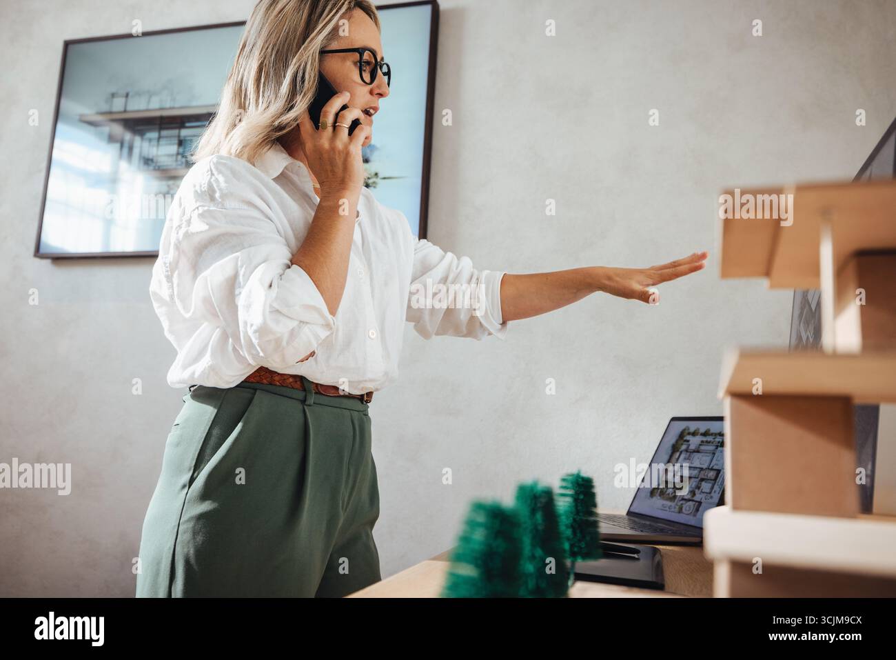 Eine professionelle Frau analysiert ein Miniaturmodell, während sie in einem Studio telefoniert. Stockfoto