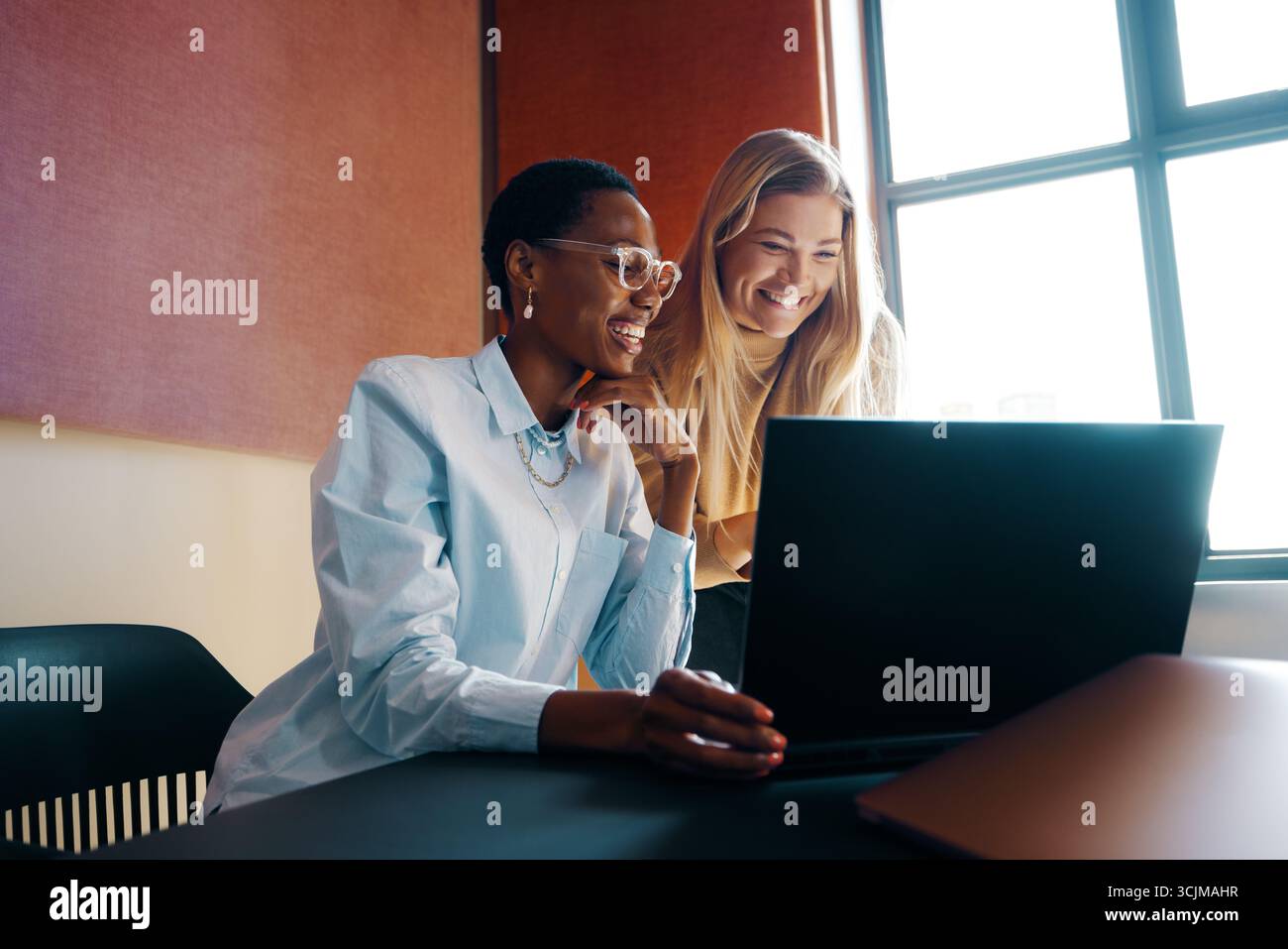 Zwei junge Frauen, eine Afrikanerin und eine kaukasierin, arbeiten zusammen und lächeln, während sie in einer modernen Büroumgebung Ideen austauschen. Stockfoto