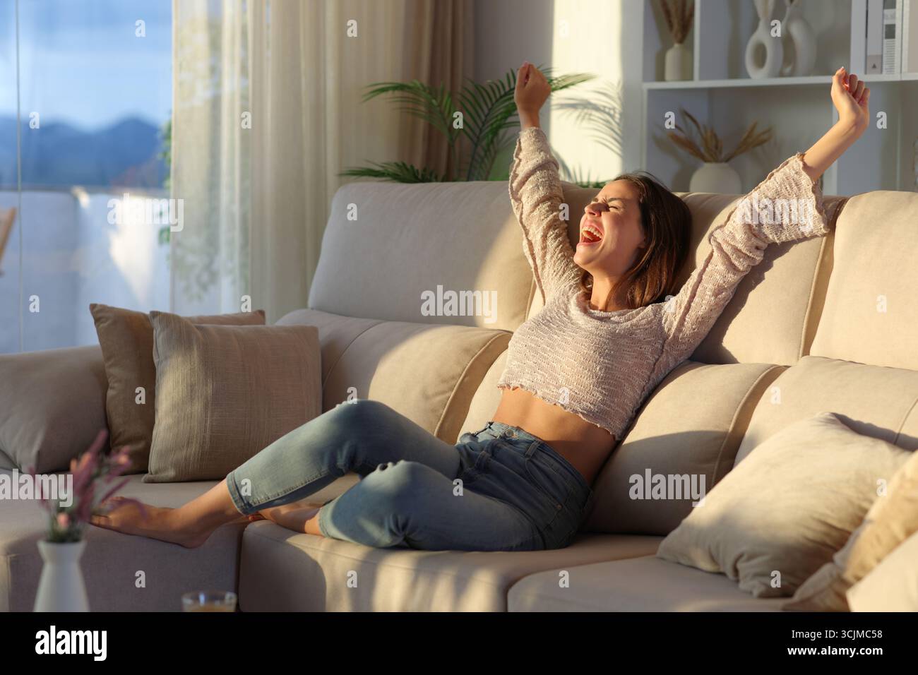Aufgeregte Frau zu Hause, die den guten Tag feiert und auf einer Couch die Arme hebt Stockfoto