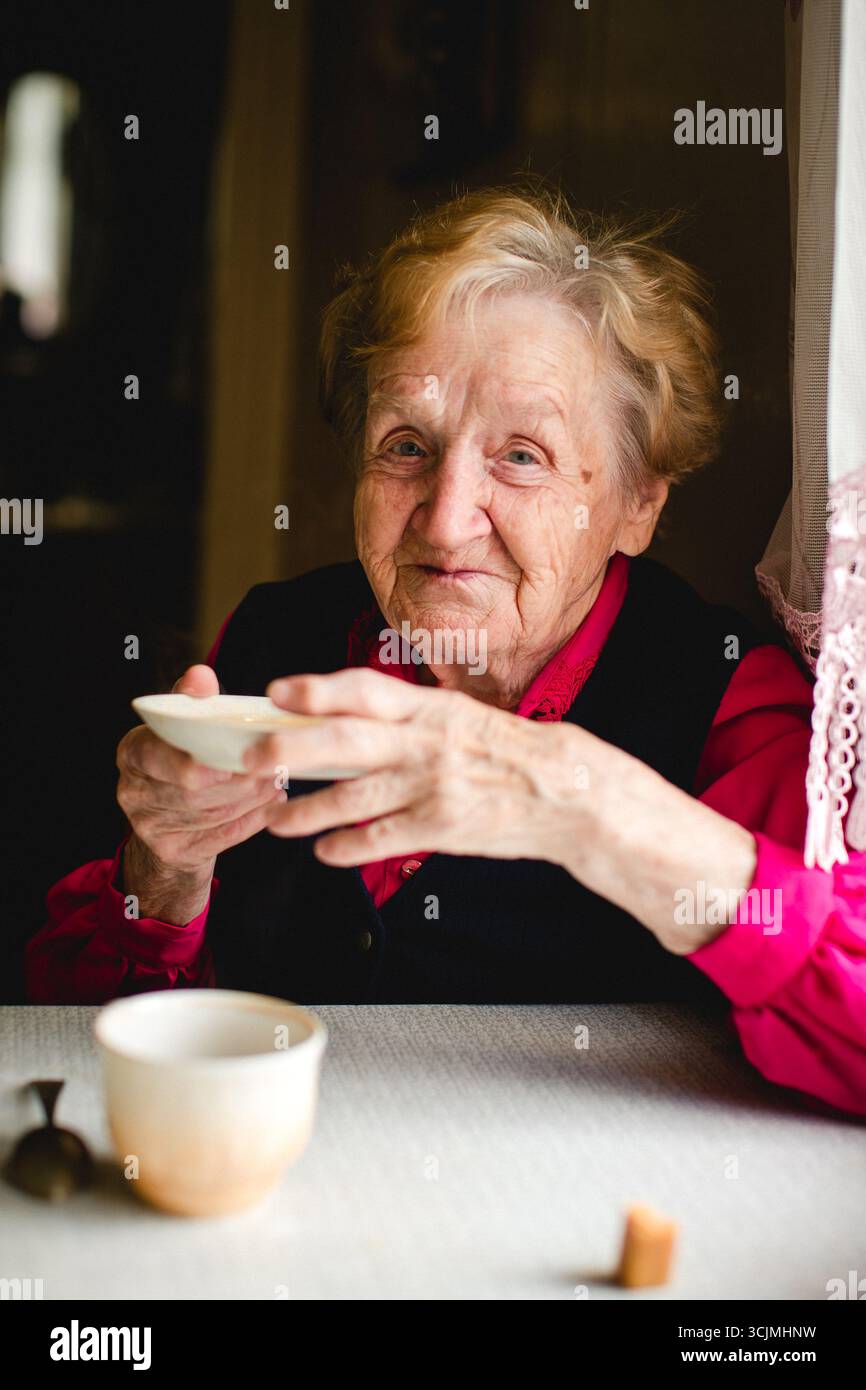 Eine ältere Frau sitzt an einem Tisch und trinkt Tee aus einer Untertasse. Das Porträt strahlt Einfachheit, Tradition und die stille Anmut des Alltags aus. Stockfoto