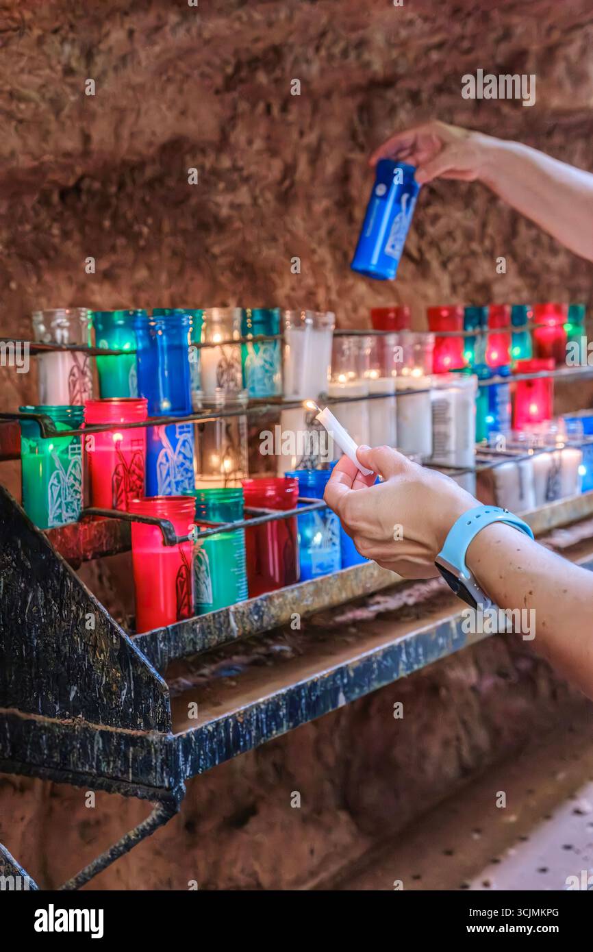 Person beleuchtet farbenfrohe Votivkerzen im historischen religiösen Höhlenschrein. Traditionelles Gebetsritual, spirituelle Hingabe, katholische Klostertradition Stockfoto