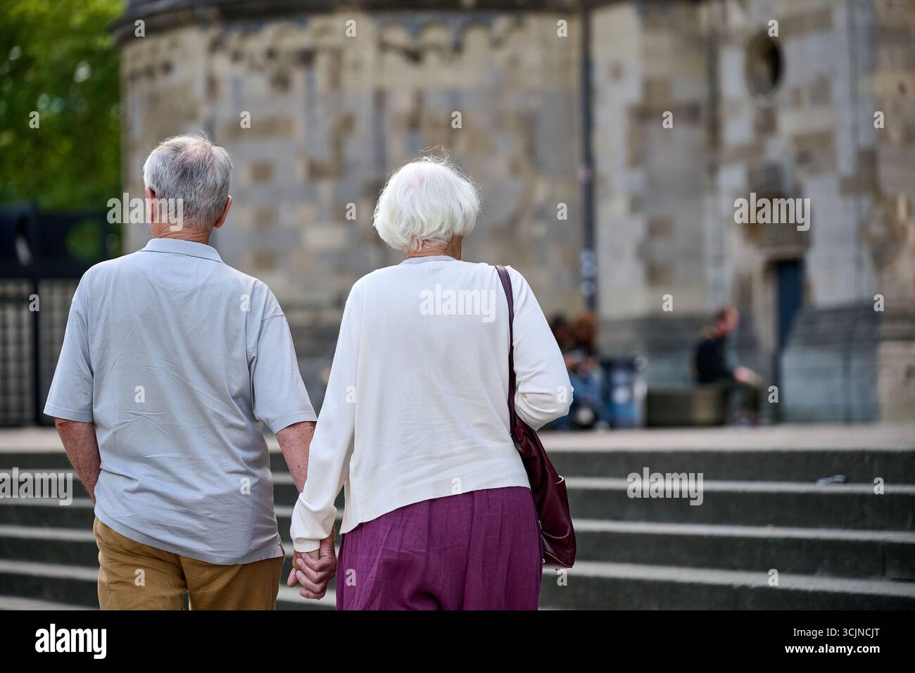 Seniorenpaar, die auf dem Bürgersteig der plaza in Richtung ferne Architektur, Tageslicht, offener horizontaler Rahmen für Lifestyle, Reisen und tägliches aktives Leben gehen Stockfoto