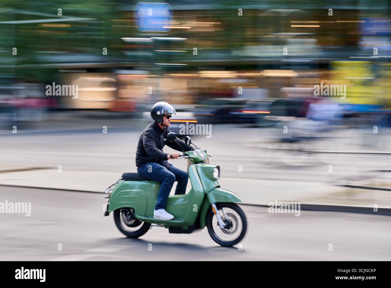 Mann auf grünem Roller in der Stadtstraße, starke Bewegungsunschärfe mit aktiver Bewegung, horizontale Actionaufnahme bei Tageslicht für urbane Mobilität, Lifestyle, an Stockfoto