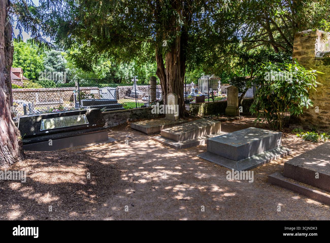 Der Familienfriedhof des George-Sand-Hauses (Maison de George Sand) in Nohant, Gemeinde Nohant-Vic, im Berry, Departement Indre, Centre-Val de Loire, Frankreich, liegt im Schatten einer alten Eiche. Steintombs und Grabplatten stehen auf sandigem Boden. Ein dunkles Grab mit einer Metallplakette steht links, und schlichte Grabsteine stehen in der Mitte. Eine Schuttsteinmauer grenzt den Friedhof rechts ab, vor der ein Rhododendron wächst. Im Hintergrund trennt ein Drahtgitterzaun den Familienfriedhof vom Dorffriedhof mit seinen Kreuzen und Denkmälern. Die Schriftstellerin starb am 8. Juni 18 Stockfoto