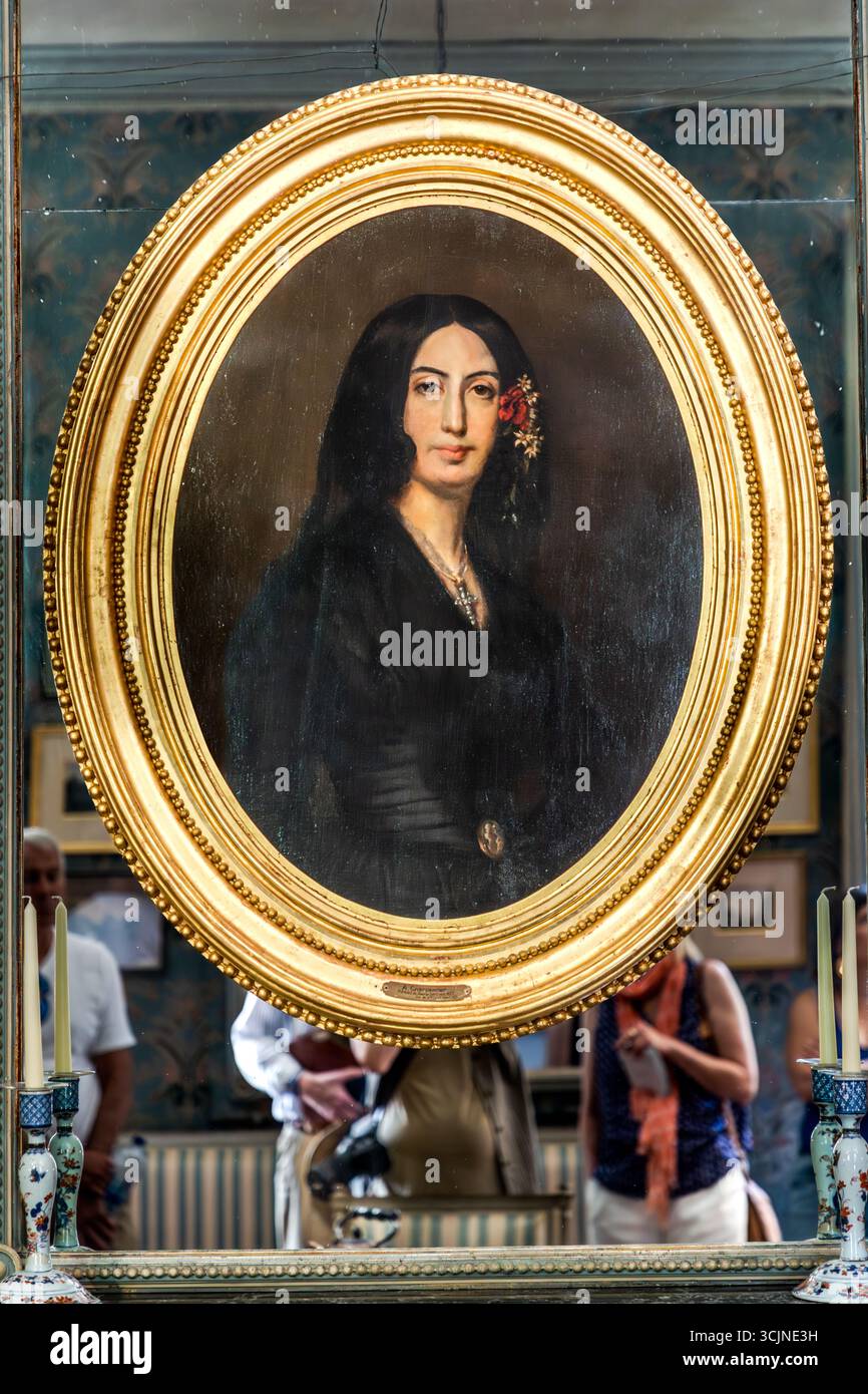 Ein ovales Porträt der jungen George Sand von Auguste Charpentier, datiert 1837, hängt im Salon des George-Sand-Hauses (Maison de George Sand) am Place Sainte-Anne in Nohant, Gemeinde Nohant-Vic, im Berry, Département Indre, Centre-Val de Loire, Frankreich. Das Gemälde zeigt die Schriftstellerin mit langem schwarzem Haar, einer roten Blume im Haar, einem schwarzen Kleid und einem Kreuz an einer Kette. Der vergoldete Perlenrahmen trägt eine kleine Namensplakette. Das Bild hängt vor einem Spiegel, der Besucher auf der Führung reflektiert, mit Porzellanleuchtern auf beiden Seiten. Im Salon steht eine Kopie, und der Ursprung Stockfoto