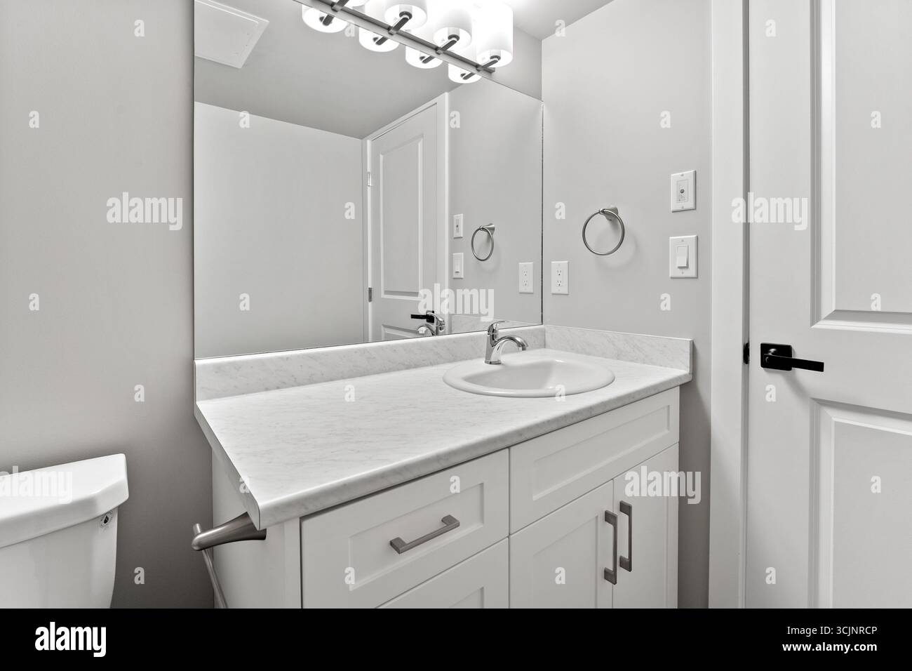 Badezimmer mit weißem Waschbecken und Toilette. Das Bad ist sauber und gut beleuchtet. Der Spiegel ist groß und reflektiert das Licht der Deckenleuchten Stockfoto