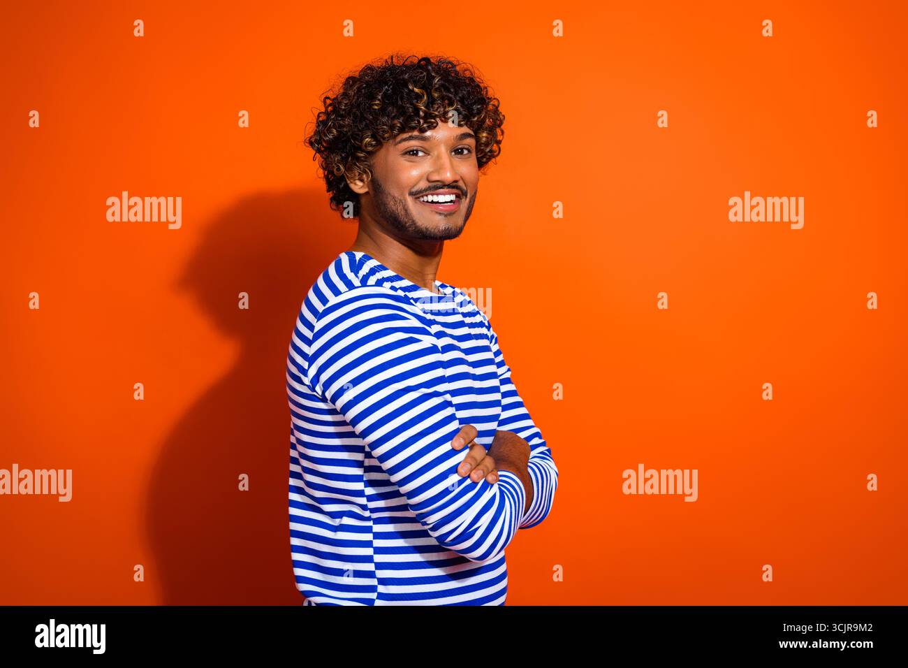 Junger Mann in einem gestreiften, von Seemännern inspirierten Hemd posiert vor einem orangen Hintergrund mit einer fröhlichen und selbstbewussten Einstellung Stockfoto