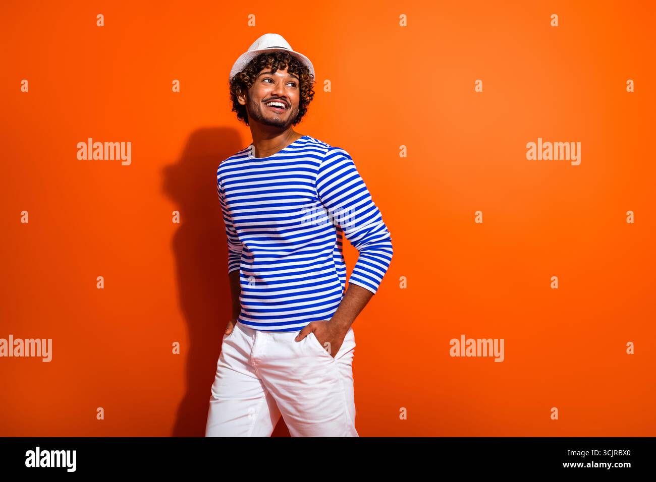 Positiver, stylischer Mann im gestreiften Matrosen-Outfit, der vor leuchtend orangefarbenem Hintergrund posiert und Sommermode und Reisen repräsentiert Stockfoto