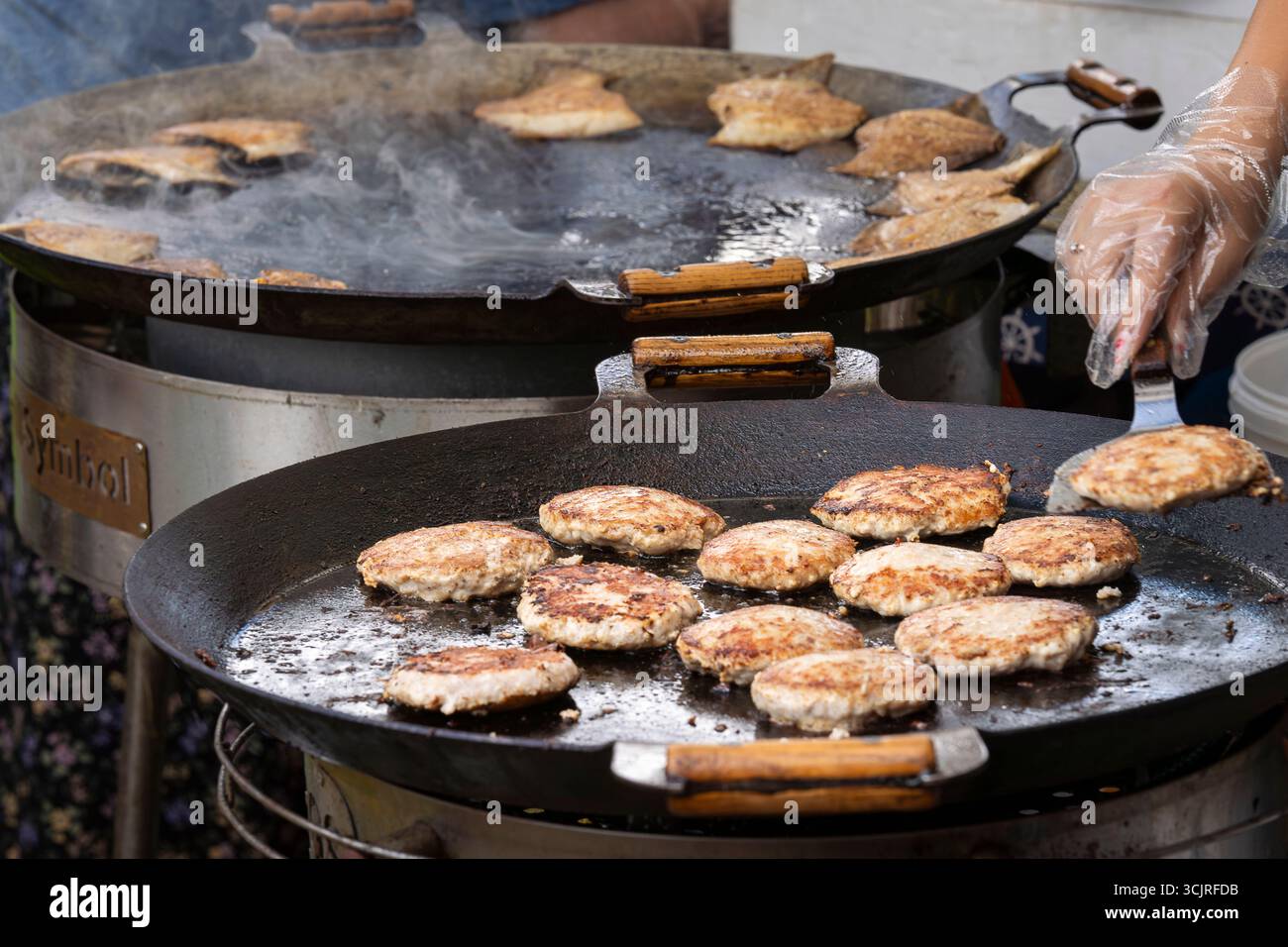 Auf einem großen runden Grill werden Fischpatties gebraten. Frisches Fischkotelett in einem Bratpfannengrill. Grillfest im Stadtpark. Street Fast Food. Stockfoto