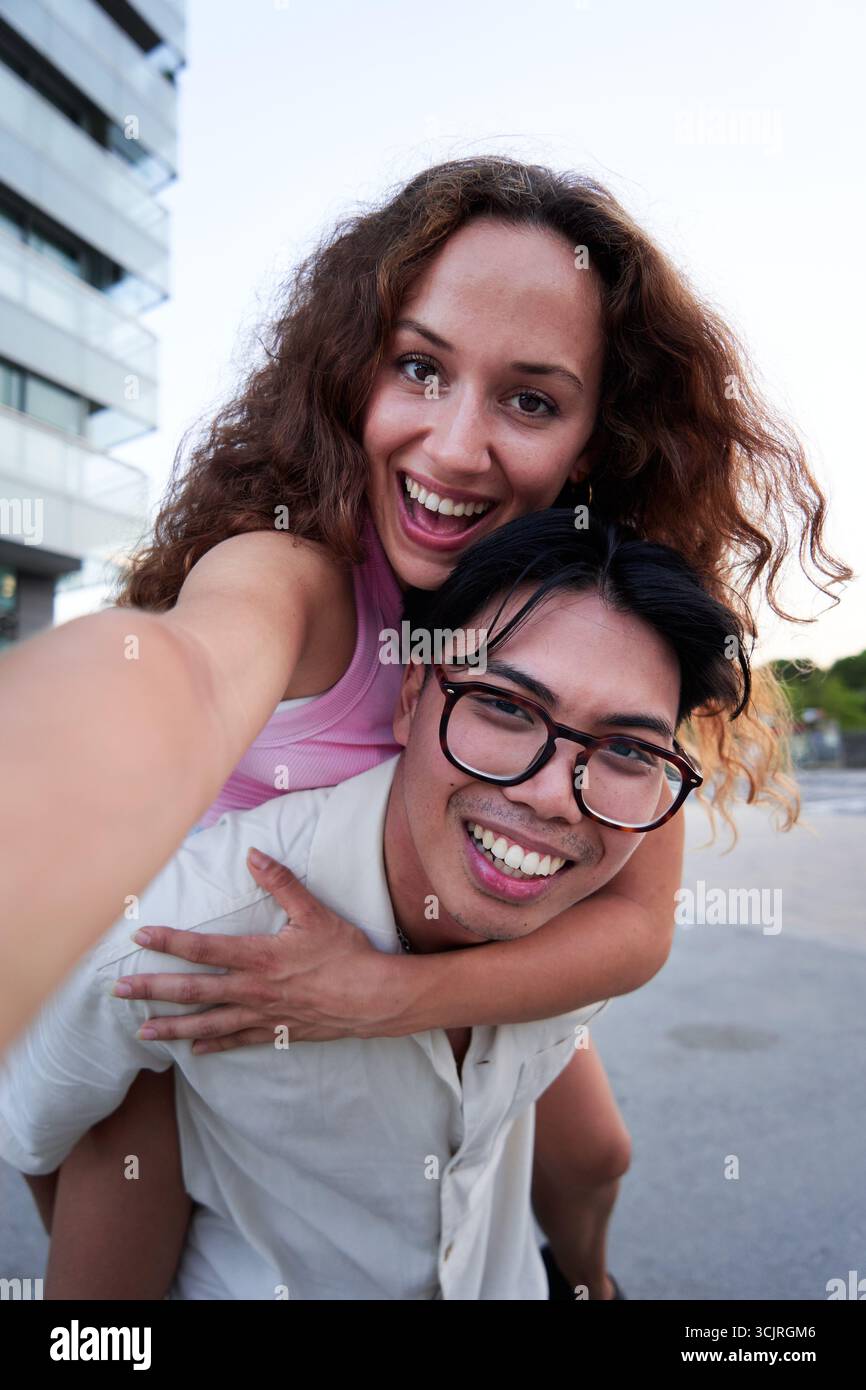 Selfie eines jungen Paares, das in die Kamera schaut, während der Freund seiner Freundin Huckepack gibt. Stockfoto