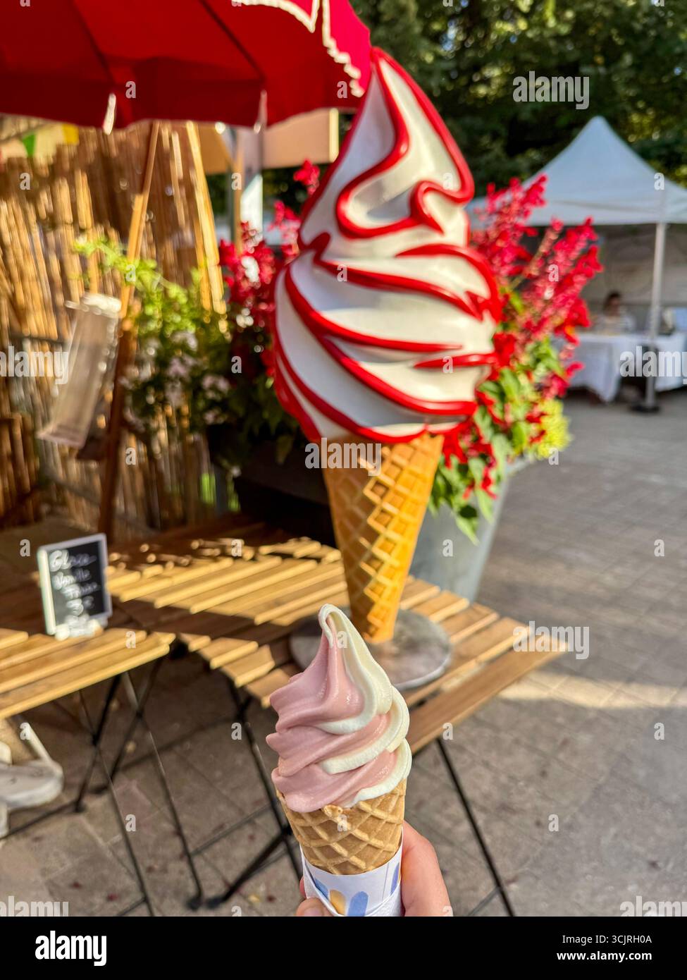 Person, die eine Erdbeervanilleeiskegel vor einem großen dekorativen Eismodell hält, im Hintergrund verschwimmt - Smartphone-aufgenommenes Stockfoto