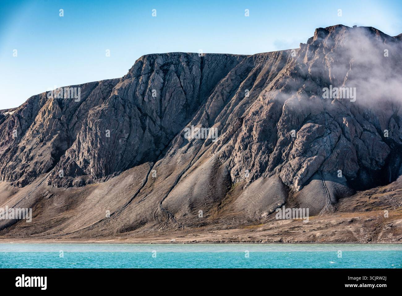 FAKSEVAGEN SVALBARD NORWEGEN ARKTISCHE LANDSCHAFT // SPITZBERGEN, Svalbard und Jan Mayen — entlang der Küste Spitzbergens, der größten Insel des Svalbard Archipels, ist Eine zerklüftete, bergige Landschaft zu sehen. Die steilen, felsigen Hänge sind charakteristisch für das arktische Gelände, wobei Geröllhänge und Rinnen die Berghänge markieren. Türkisfarbenes Wasser, wahrscheinlich der Arktische Ozean, läuft am Fuß der Berge. Nebelschwaden ziehen über die oberen Bereiche der Gipfel und tragen zur dramatischen Atmosphäre dieser abgelegenen Polarregion bei. Svalbard ist bekannt für seine Gletscher, Tundra und üppigen Wildlif Stockfoto