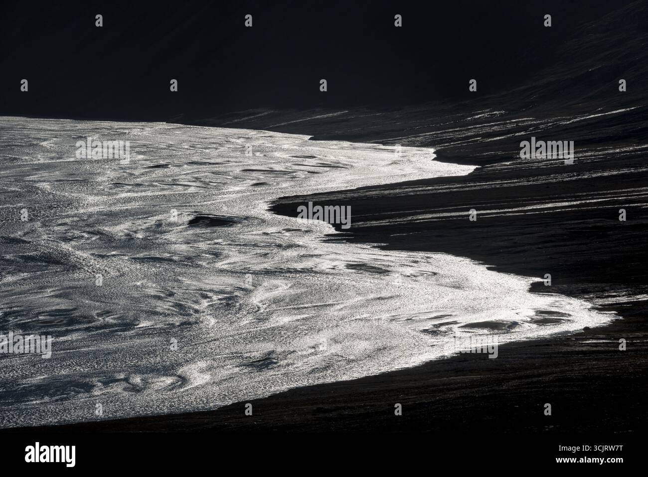 Faksevagen Wasser glitzert im Sonnenlicht Svalbard // SVALBARD UND JAN MAYEN — die Sonne scheint von der Wasseroberfläche, während sie an einem dunklen, steinigen Ufer in Svalbard entlang fließt. Diese raue Landschaft, die durch das Zusammenspiel von Licht und Schatten auf Wasser und Land gekennzeichnet ist, ist typisch für den abgelegenen arktischen Archipel. Der Titel „Faksevagen Water Glistening in Sunlight Svalbard“ deutet darauf hin, dass es sich bei diesem speziellen Ort um eine Bucht oder einen Einlass innerhalb der Region Svalbard handelt. Svalbard ist bekannt für seine dramatische natürliche Schönheit und seine extreme arktische Umgebung. Stockfoto