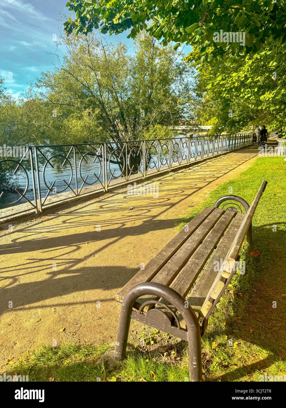 Flussweg mit Holzbank, Metallgeländer, grünen Bäumen und ruhigem Wasser unter teilweise bewölktem Himmel, friedliche Parkszene mit Spaziergängen. Stockfoto