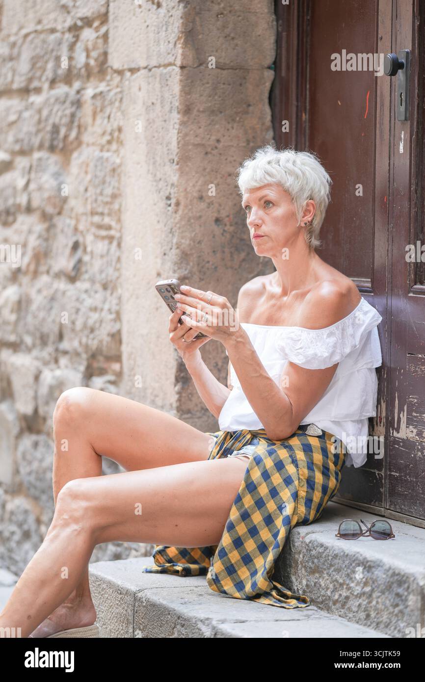 Eine Frau in einem weißen Hemd und einem gelb-schwarzen karierten Rock sitzt auf einer Stufe. Sie schaut sich ihr Handy an Stockfoto