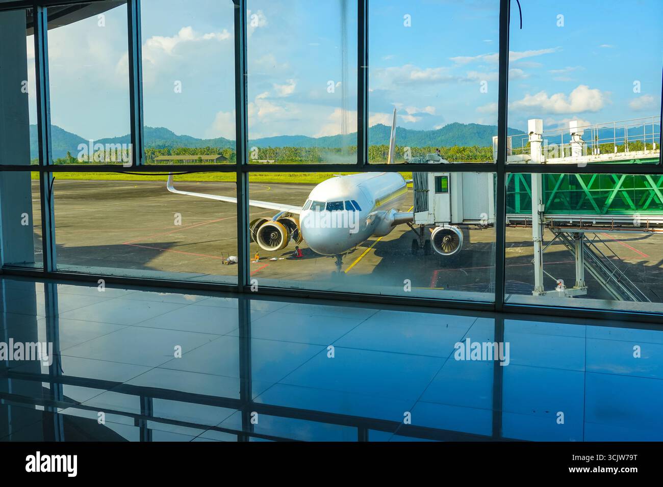 Ein modernes Flugzeug sitzt am Gate, das durch das Fenster des Flughafenterminals sichtbar ist, mit einer reflektierenden Bodenfläche. Diese Szene ist ideal für Reisen oder aviat Stockfoto