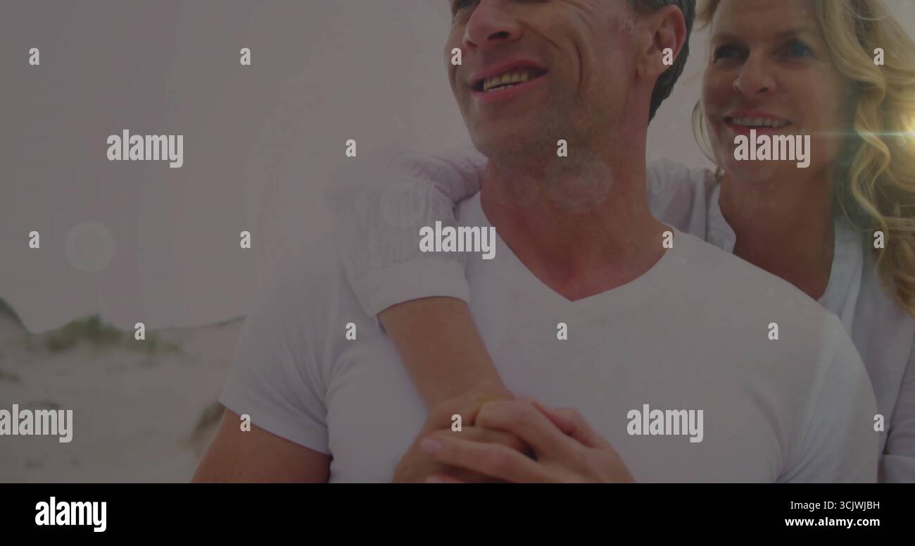 Lächelndes, reifes Paar umarmt am Sandstrand mit grasbewachsenen Dünen, Ring sichtbar auf der linken Hand der Frau Stockfoto