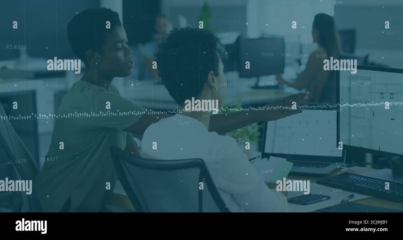 Zwei Kollegen, die sich über den Schreibtisch beugen und im Büro Daten auf zwei Monitoren mit Dokumenten überprüfen Stockfoto
